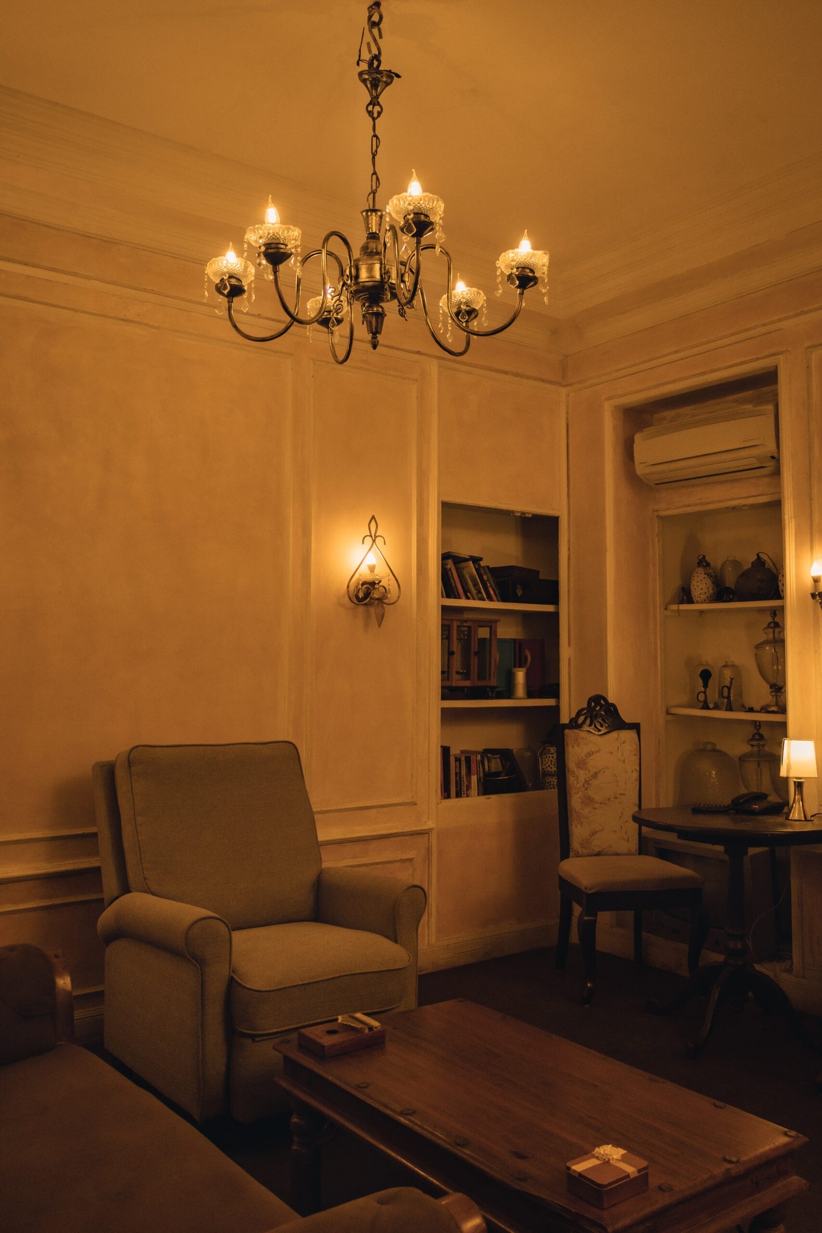 The lounge in the evening: a brass chandelier, panelled walls, a recliner and a carved wooden chair beside a low table