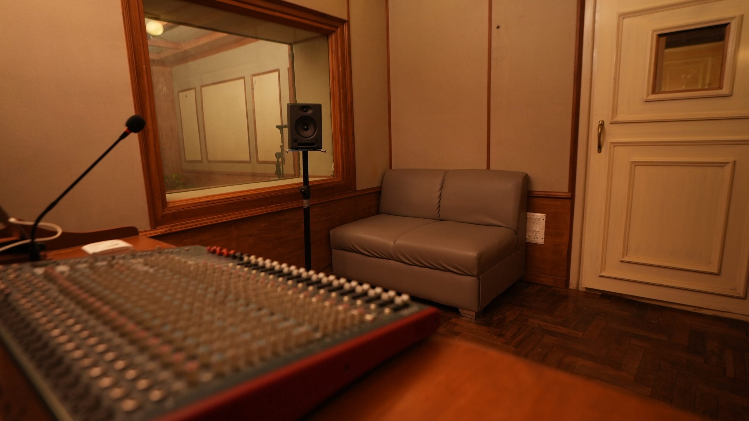 A mixing console in the foreground with the booth window and a monitor speaker behind